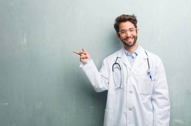 junger freundlicher Arzt vor einer Grunge-Wand mit einem Kopierraum, der zur Seite zeigt, lächelnd überrascht etwas präsentiert, natürlich und lässig - Foto, Bild
