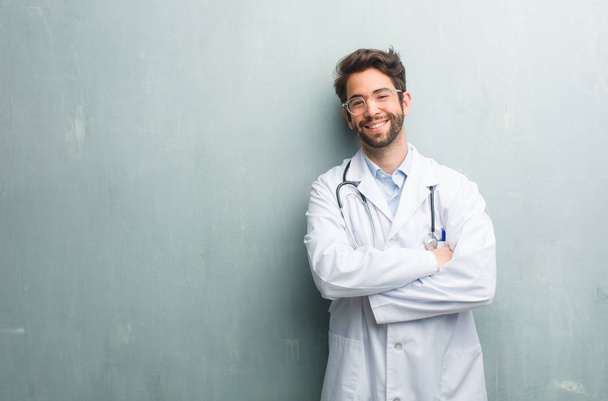 Young friendly doctor man against a grunge wall with a copy space crossing his arms, smiling and happy, being confident and friendly - Photo, Image