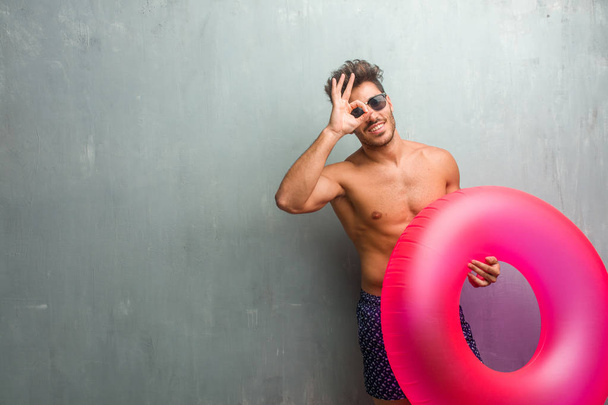 Young athletic man wearing a swimsuit against a grunge wall cheerful and confident doing ok gesture, excited and screaming, concept of approval and success - Valokuva, kuva
