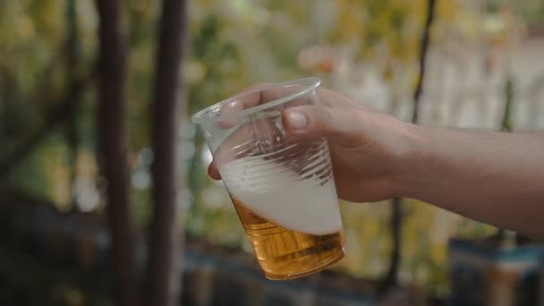 Mann hält Hand und schüttelt langsam einen durchsichtigen Plastikbecher mit leichtem Bier  - Filmmaterial, Video