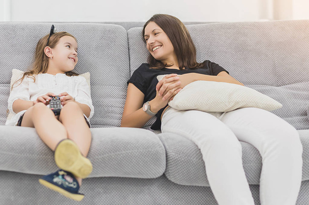 Mother and daughter are having a lot of fun while watching TV in the living room sitting on the couch - Fotó, kép