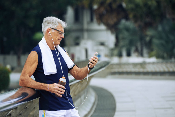 Terve vanhempi mies tarkistaa älypuhelimen harjoittelun jälkeen ulkona
 - Valokuva, kuva