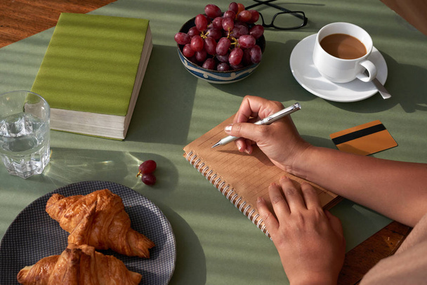 Unrecognizable woman sitting at kitchen table and taking necessary notes, appetizing croissants and cup of fragrant coffee in cup - Photo, Image