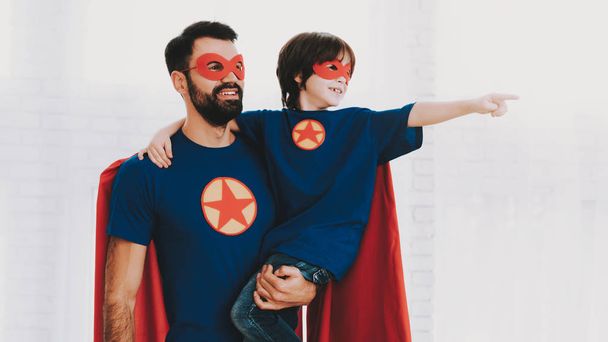 Father And Son. Red And Blue Superhero Suits. Masks And Raincoats. Posing In A Bright Room. Young Happy Family Holiday Concept. Resting Together. Save The World. Get Ready. Strong And Powerful. - Foto, immagini