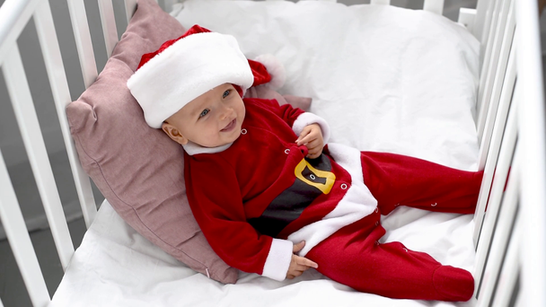 очаровательный улыбающийся ребенок в костюме Санты, лежащий на подушке в кроватке дома, рождественская концепция
 - Кадры, видео
