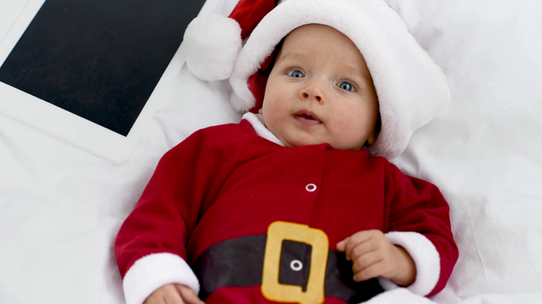 upset adorable baby in santa costume lying with tablet in crib at home, christmas concept - Footage, Video