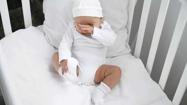 schattig baby in witte kleren zitten in de wieg en wrijven oog thuis - Video