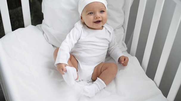 ihana vauva valkoisissa vaatteissa istuu pinnasängyssä ja hymyilee kotona
 - Materiaali, video