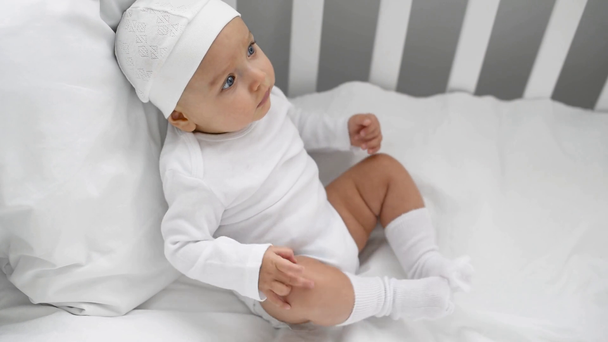 adorable infant in white clothes sitting in crib and moving legs at home - Filmati, video