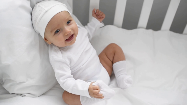 ευτυχισμένος αξιολάτρευτο μωρό με λευκά ρούχα που κάθεται στο παχνί στο σπίτι - Πλάνα, βίντεο