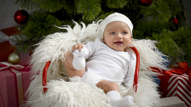 adorable sonriente bebé sentado en silla cerca de árbol de Navidad en casa
 - Metraje, vídeo