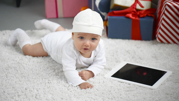 schattig ongelukkig baby liggend op witte tapijt in de buurt van tablet met leeg scherm thuis, Kerstmis concept - Video
