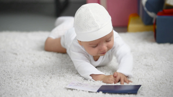 adorable baby lying on white carpet and touching passport with ticket at home, christmas concept - Filmati, video