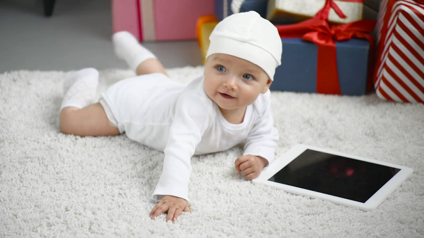 lachende schattig baby liggend op witte tapijt in de buurt van tablet met leeg scherm thuis, Kerstmis concept - Video