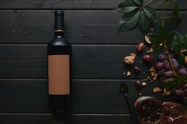 κάτοψη του μπουκάλι κρασί με την ετικέτα κενή, σταφύλια, καρύδια και μαρμελάδα σε ξύλινη επιφάνεια - Φωτογραφία, εικόνα
