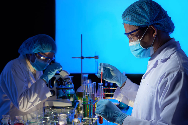 Asiatische Wissenschaftler mit Gummihandschuhen und Schutzmasken arbeiten im dunklen Labor zusammen - Foto, Bild