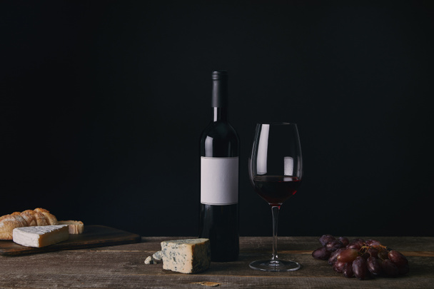 bottle of wine with blank label, glass of red wine, cheese and grapes on wooden table - Fotó, kép