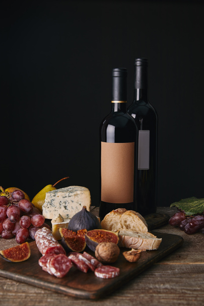 close-up view of bottles of red wine, fruits and delicious cheese with salami on wooden table   - Foto, imagen