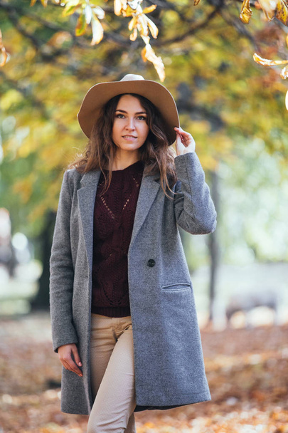 Portrait of beautiful young woman walking outdoors in autumn park in cozy coat and hat. Warm sunny weather. Fall concept. - Photo, Image