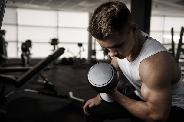 Handsome man doing biceps lifting with dumbbell on bench in a gym - Photo, Image