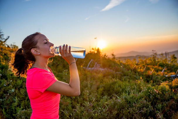 Woman drinking water from the bottle after running training. Young woman cross running in the mountains. - Foto, afbeelding