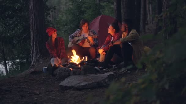 Cheerful travelers are singing songs and playing the guitar sitting around fire in forest in the evening and having fun with beautiful nature around. People and music concept. - Footage, Video