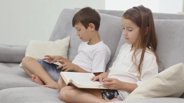 little cute boy play tablet while his sister reads a book on sofa at home - Materiał filmowy, wideo