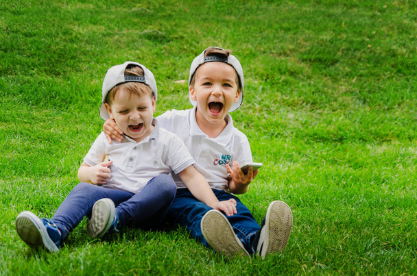Два мальчика трех лет в белых футболках, джинсах и кепках сидят на траве и обнимаются. Два близких друга, два кузена. Семейные отношения между братьями
 - Фото, изображение