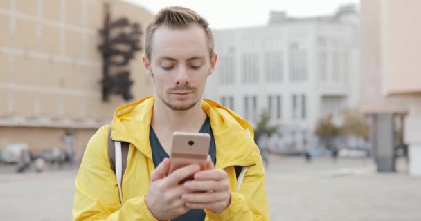 Hombre caucásico en chaqueta amarilla usando mensajes de texto de teléfonos inteligentes navegar mensajes de un teléfono móvil, comunicarse en la ciudad
 - Metraje, vídeo