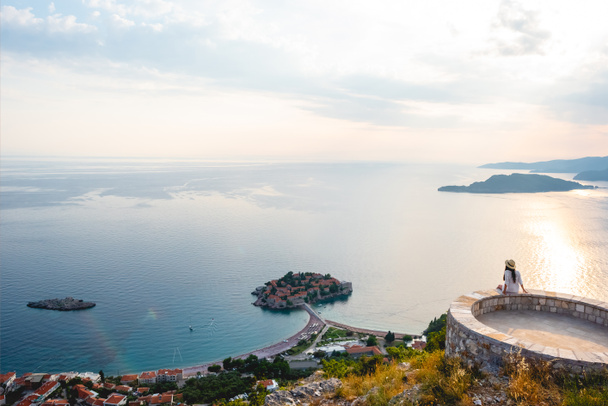 woman sitting on viewpoint and looking at island of Sveti Stefan during sunset in Budva, Montenegro - Photo, Image