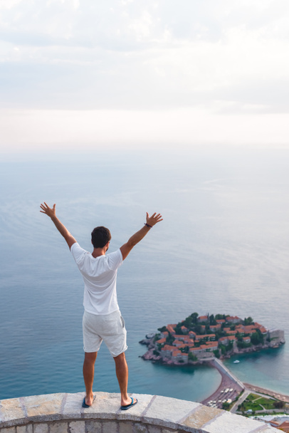 back view of man with raised hands looking at island of Sveti Stefan with hotel resort in Adriatic sea, Budva, Montenegro - Photo, Image