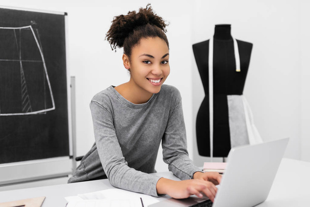 Uśmiechnięta i pozytywna kobieta z piękną twarzą i fryzurą przy użyciu laptopa, projekt pracy w miejscu pracy. Portret Afroamerykańskiego projektanta siedzącego w biurze na poddaszu. Udane interesy. Kariera - Zdjęcie, obraz