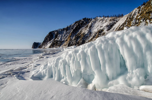 Lake Baikal is covered with ice and snow, strong cold, thick clear blue ice. Icicles hang from the rocks. Lake Baikal is a frosty winter day. Amazing place - 写真・画像