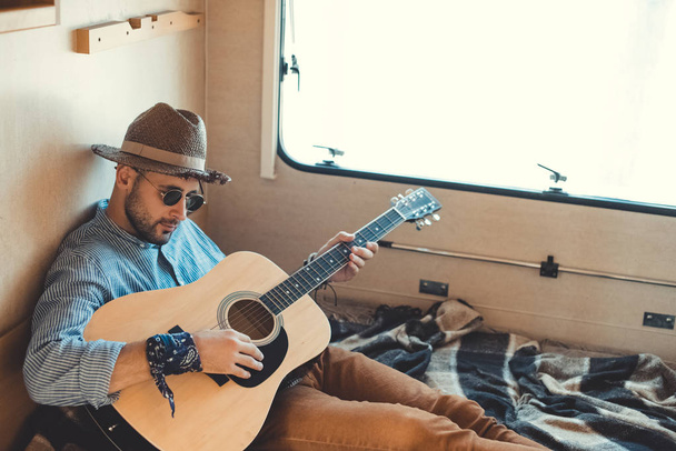 handsome man in hat playing guitar inside of campervan - Foto, afbeelding