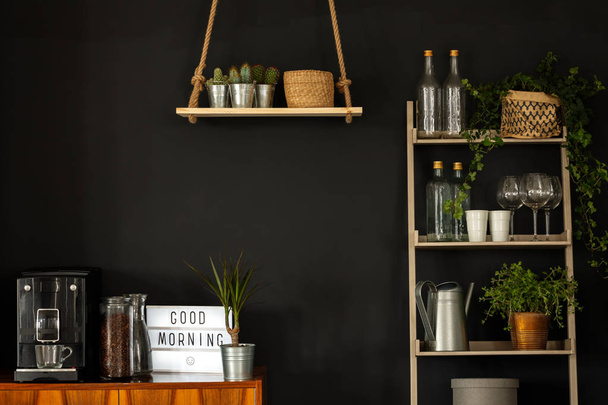 Πραγματική φωτογραφία ενός εσωτερικού μοντέρνα κουζίνα με ράφια, φυτά, μηχανή καφέ και ποτήρια κρασιού σε ένα μαύρο τοίχο - Φωτογραφία, εικόνα