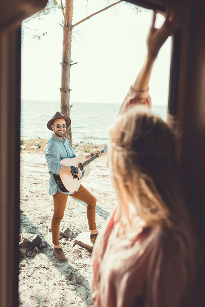 selective focus of girl standing in door of campervan while man playing guitar - Zdjęcie, obraz