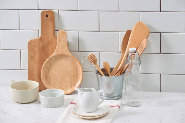 Simple rustic kitchenware against white wooden wall: rough ceramic pot with wooden cooking utensil set, stacks of ceramic bowls, jug and wooden trays. - Photo, image
