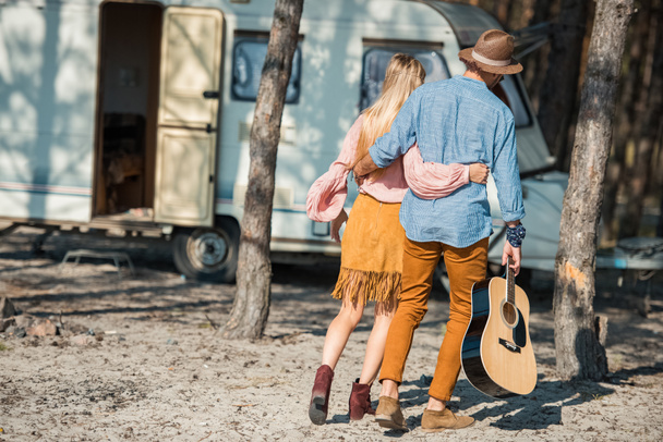 rear view of hippie couple hugging and walking with guitar near trailer - Φωτογραφία, εικόνα