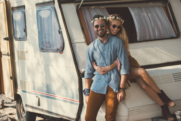 happy young couple in wreaths and sunglasses hugging and sitting on campervan   - Zdjęcie, obraz