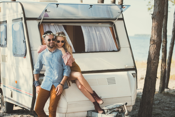 hippie couple in wreaths and sunglasses hugging and sitting on trailer near sea - Foto, imagen