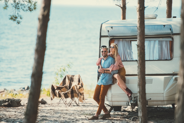 happy hippie couple in wreaths hugging and sitting on campervan near sea - Foto, Bild