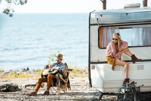 beautiful hippie girl sitting on campervan while man playing guitar near sea - Photo, Image