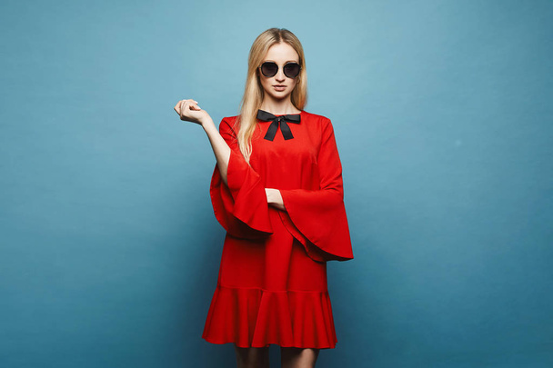 Fashionable and sexy blonde model girl in stylish sunglasses and in glamorous short red dress with black bow posing at blue background in studio, isolated - Photo, image