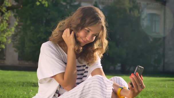 Mladý, usměvavý, Kavkazský bruneta sedí v parku na trávě, pomocí chytrého telefonu, takže selfie, univerzita v pozadí - Záběry, video