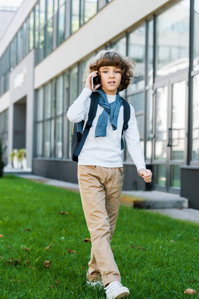 full length view of boy with backpack talking by smartphone and looking at camera - Fotografie, Obrázek