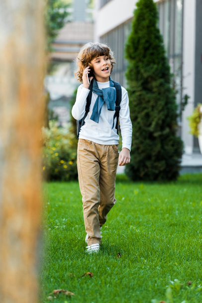selective focus of smiling boy with backpack talking by smartphone and walking on green lawn - Foto, immagini