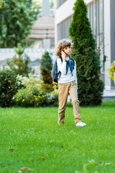 full length view of preteen schoolboy with backpack talking by smartphone and walking on green lawn - Foto, Bild