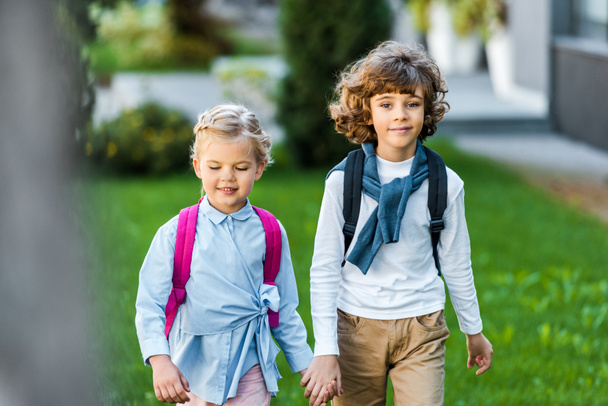 adorable happy little schoolkids holding hands and walking on green lawn - Photo, Image