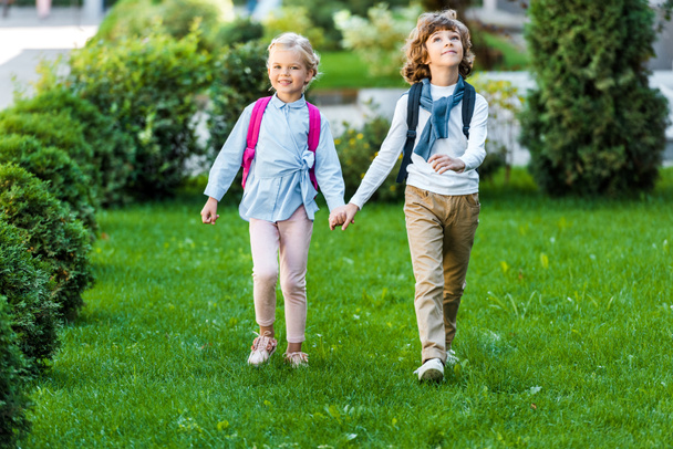 full length view of adorable happy schoolkids with backpacks holding hands and walking on green lawn - Photo, Image