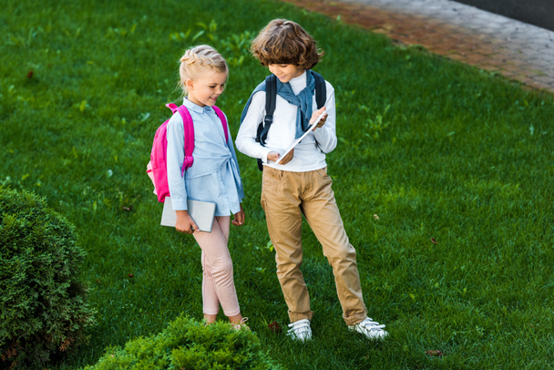 wysoki kąt widzenia dzieci z plecakami, trzymając cyfrowego tabletu stojąc razem na zielony trawnik  - Zdjęcie, obraz